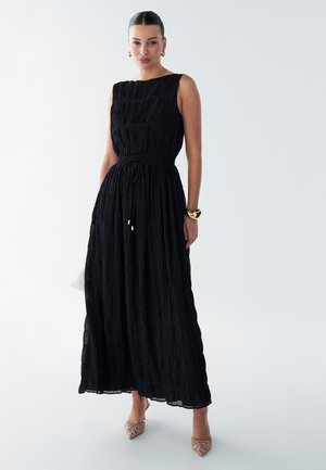 Black maxi dress with pleated texture, sleeveless design, elastic waist, and a flowing skirt. Worn with beige embellished heels.