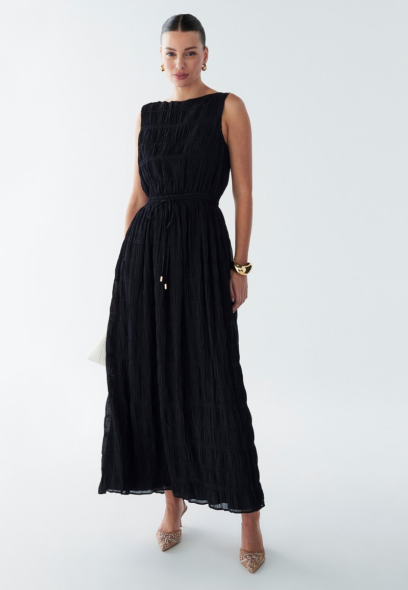 Schwarzes Maxi-Kleid mit gefalteter Textur, ärmellosem Design, elastischem Bund und fließendem Rock. Getragen mit beigefarbenen, verzierten Sneakers.