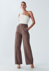 Biały prążkowany top na jedno ramię w połączeniu z wysokim stanem, brązowymi spodniami o szerokich nogawkach, ozdobionymi plisami i kieszeniami bocznymi. Sandały w neutralnym kolorze.