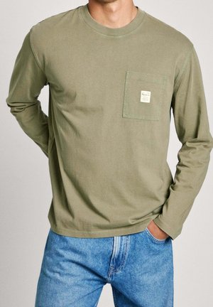 Mann trägt ein olivgrünes Langarmhemd mit Brusttasche und blaue Jeans, steht mit einer Hand in der Tasche und der anderen hinter dem Rücken.
