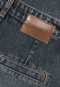 Tmavomodré džíny vyrobené z denimu s textúrovaným povrchom, oranžovým prešívaním a hnedou koženou nášivkou s embosovaným nápisom "LAMANIA."