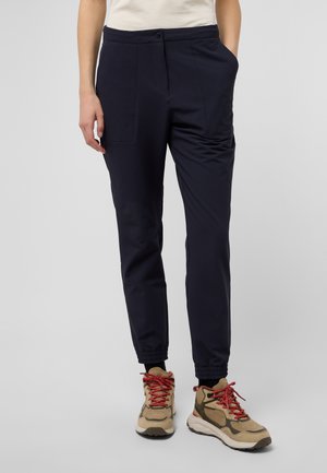 Marineblaue, tapered Hose mit Knopfverschluss, Seitentaschen und elastischen Bündchen. Kombiniert mit beigen und braunen Wanderschuhen mit roten Schnürsenkeln.
