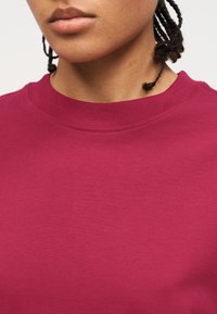 Άτομο με πλεξούδες που φοράει jednounto μπλουζάκι στρογγυλής λαιμόκοψης σε βαθύ ροζ χρώμα, εμφανίζοντας τον λαιμό και το κάτω μέρος του προσώπου χωρίς ορατή έκφραση.