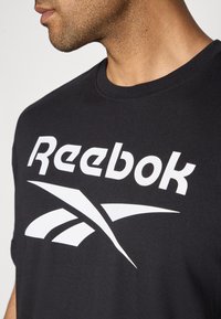 T-shirt noir en coton arborant un logo "Reebok" blanc bien visible et des éléments de design dynamiques. Gros plan sur le col et une partie de l'épaule.
