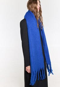 Blød blå uldsjaal med en lang, rektangulær form og frynser i kanten, draperet om halsen på en person iført en sort frakke.