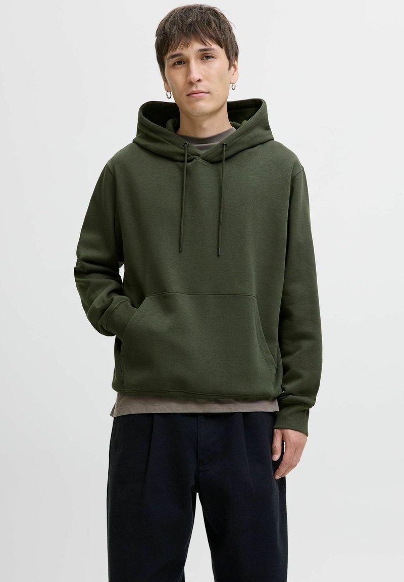 Olive green hoodie made of soft fabric with a front pocket, drawstring hood, and ribbed cuffs, worn over a gray shirt and dark pants.