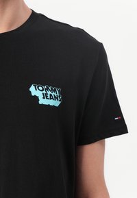 Black cotton T-shirt with a light blue "TOMMY JEANS" logo on the chest and a small red, white, and blue accent on the sleeve.