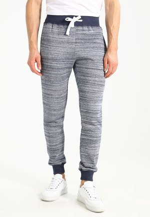 Uomo che indossa pantaloni jogger grigi con coulisse, vita e polsini blu navy, abbinati a sneakers bianche e camicia bianca.