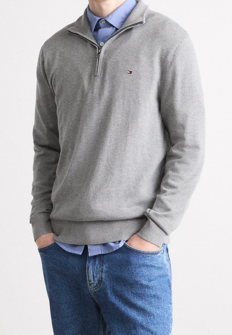 Maglione grigio con collo a mezza zip, texture a maglia e polsi a costine. Camicia blu indossata sotto e abbinata a jeans blu.