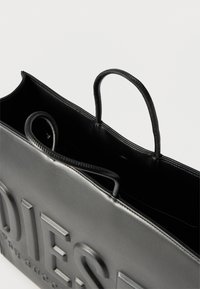 Μαύρη τσάντα χειρός από δέρμα με ανάγλυφο μεγάλο λογότυπο μπροστά και δύο λουράκια, πάνω σε λευκό φόντο.