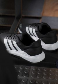 Paire de chaussures de sport noires avec des rayures blanches et des semelles, posées sur un banc sombre avec un sol texturé en dessous.