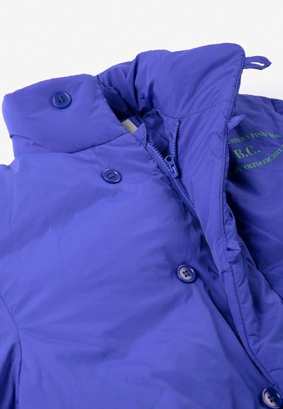Veste matelassée violette avec fermetures par boutons et zip ainsi qu'un col haut, montrant une partie du texte vert imprimé à l'intérieur.