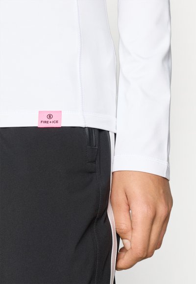 Λευκή μπλούζα με μακριά μανίκια και επίπεδες ραφές, με ροζ ετικέτα που γράφει "FIRE+ICE", σε συνδυασμό με μαύρα παντελόνια που διαθέτουν τσέπη με φερμουάρ στο πλάι.