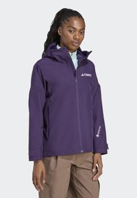 Ljubičasta vodootporna jakna s kapuljačom, koja ima Terrex logotip. Uključuje Gore-Tex oznaku i džepove sa patentnim zatvaračem na prednjem dijelu.
