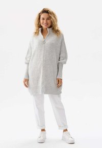 Długi, jasnoszary sweter dzianinowy z półzamkiem, żebrowanymi mankietami i zaokrąglonym dołem. Noszony na białe spodnie i białe trampki.