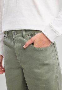 Pantalones de algodón verde oliva con un corte estándar, que cuentan con bolsillos frontales y herrajes de botón de metal. La tela tiene un acabado ligeramente texturizado.