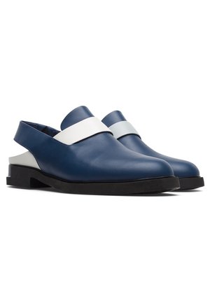 Chaussures en cuir bleu à enfiler avec des détails de sangles blanches, portées sur des pieds nus sur un fond blanc uni.