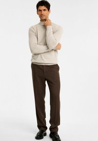 Hellbeige Rollkragenpullover, figurbetontes Design, mit langen Ärmeln. Kombiniert mit braunen, maßgeschneiderten Hosen und schwarzen Lederschuhen. Einfarbiger Hintergrund.