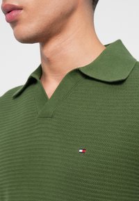 Zöld kötött polo póló, textúrált anyaggal, széles gallérral és egy kis piros, fehér és kék logóval a mellkason.
