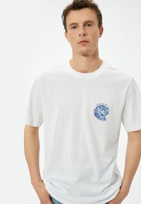 Hvid bomulds t-shirt med et blåt spiralprint på venstre bryst. Rund halsudskæring og korte ærmer, med en afslappet pasform.