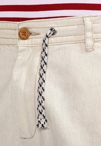 Pantalones beige con un cordón de patrón en blanco y negro, botón de madera etiquetado como "IJC Jeanscode" y fondo de tela a rayas rojas y blancas.