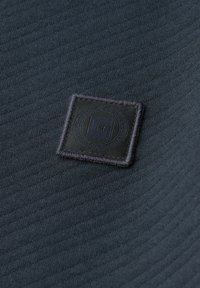 Tessuto nero con texture rigata, caratterizzato da un quadrato nero applicato con un logo circolare ricamato al centro.