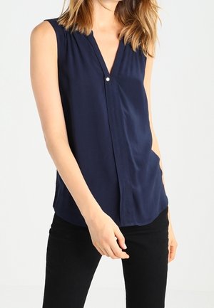 Modella che indossa una blusa senza maniche blu navy con scollatura a V e dettagli con bottoni, abbinata a pantaloni neri, in piedi contro uno sfondo chiaro.