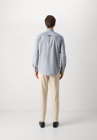 Ανοιχτό μπλε πουκάμισο με μακριά μανίκια και καμπυλωτό στρίφωμα, κουμπιά στους καρπούς και πίσω λεπτομέρεια, σε συνδυασμό με μπεζ παντελόνι και μαύρα παπούτσια.