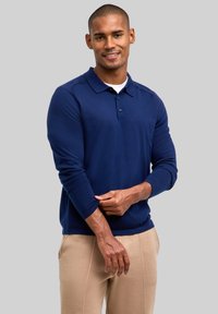 Granatowa koszulka polo z długim rękawem wykonana z gładkiej tkaniny, z klasycznym kołnierzykiem, listwą na trzy guziki oraz ściągaczami na mankietach. W połączeniu z beżowymi spodniami.
