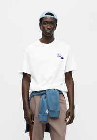 Λευκό βαμβακερό t-shirt με μπλε γραφικό σχέδιο και άνετη εφαρμογή, συνδυασμένο με καρό παντελόνι και μπλε φούτερ δεμένο στη μέση.