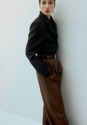 Kobieta w dużych perłowych kolczykach, czarnej kraciastej marynarce i brązowych skórzanych spodniach z wysokim stanem, stoi z rękami w kieszeniach na tle białej ściany.