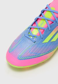 Football boot featuring a blue synthetic material with pink and green accents, textured surface, and a streamlined design with laces.