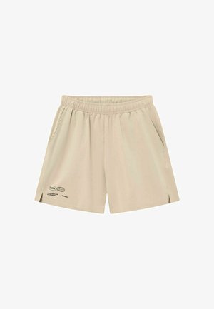 Beige shorts med elastisk talje, sidelommer og lille sort tekst samt globusgrafik nær venstre kant.