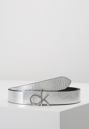 Ceinture en cuir argenté avec boucle métallique "ck", enroulée et exposée sur une surface blanche sur un fond uni.