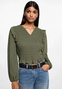 Olivgrön smockblus med volangärmar, V-ringning och kortad fåll, kombinerad med denimjeans och ett svart bälte i midjan.