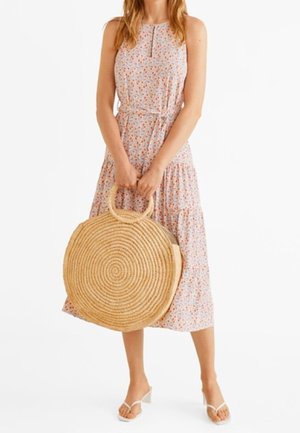 Mujer en un vestido midi sin mangas con estampado floral, sosteniendo un gran bolso de paja tejido en forma de círculo, usando sandalias de tacón blancas, de pie sobre un fondo blanco.