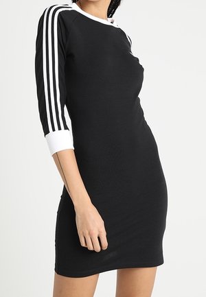 Robe noire avec des manches trois quarts, col blanc, et rayures blanches sur les manches. Fabriquée dans un tissu lisse, coupe bodycon, longueur genoux.