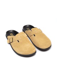Suede slip-on apavi gaismati brūnā krāsā, ar plašu siksniņu un sprādzi, melniem gumijas zoleļiem un atvērtu aizmuguri ērtai uzvilkšanai.
