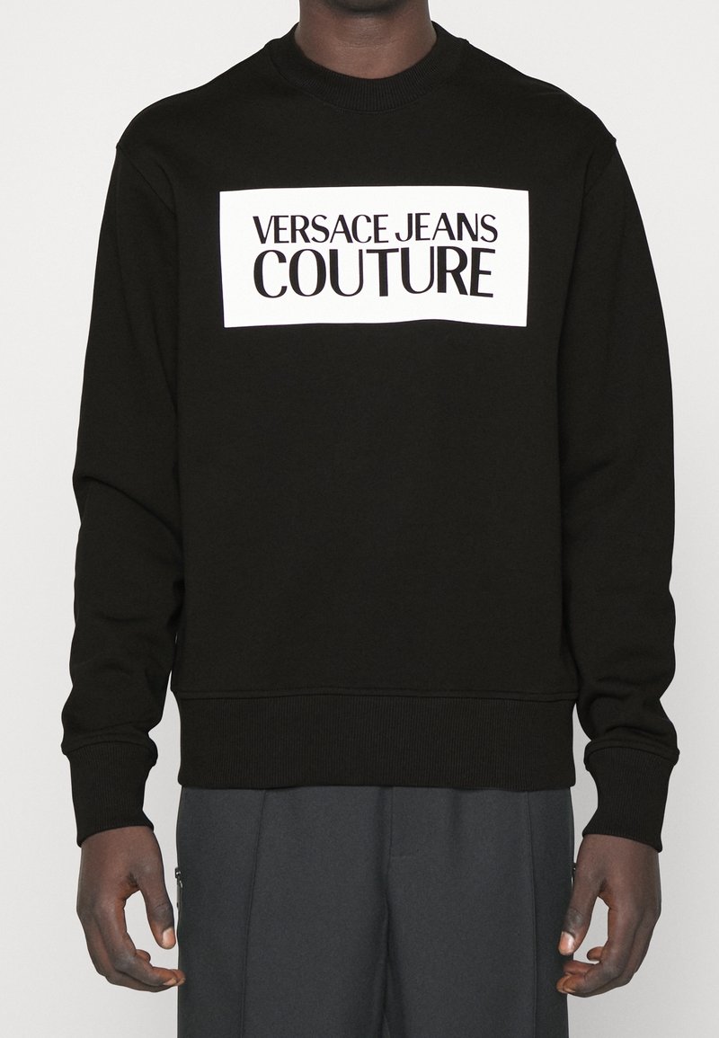 Černá mikina vyrobená z bavlny, s bílým obdélníkovým logem s nápisem "VERSACE JEANS COUTURE" tučnými černými písmeny na přední straně.