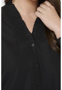 Femme portant une chemise noire boutonnée avec un col en V à volants, touchant ses cheveux près du cou et de la zone du col.
