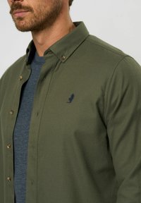 Olīvu zaļš pogu krekls ar norādītu apkakli, ar brūni dzeltenām pogu akcentiem un mazu jūras zvejnieka emblēmu uz krūtīm. Audums no kokvilnas maisījuma.