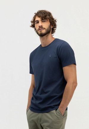 Hombre con pelo rizado y barba, vestido con una camiseta azul marino y pantalones verdes, de pie con las manos en los bolsillos frente a un fondo liso.