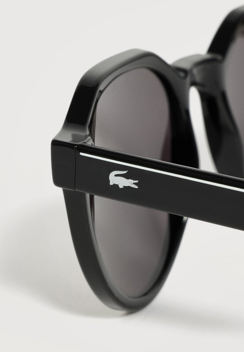 Black round sunglasses with dark lenses and a white crocodile logo on the temple arm against a light background.