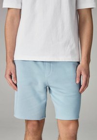 Pantaloni corti azzurri chiari realizzati in tessuto morbido, con una texture liscia, una forma aderente e una singola tasca frontale. Abbinati a una maglietta bianca.