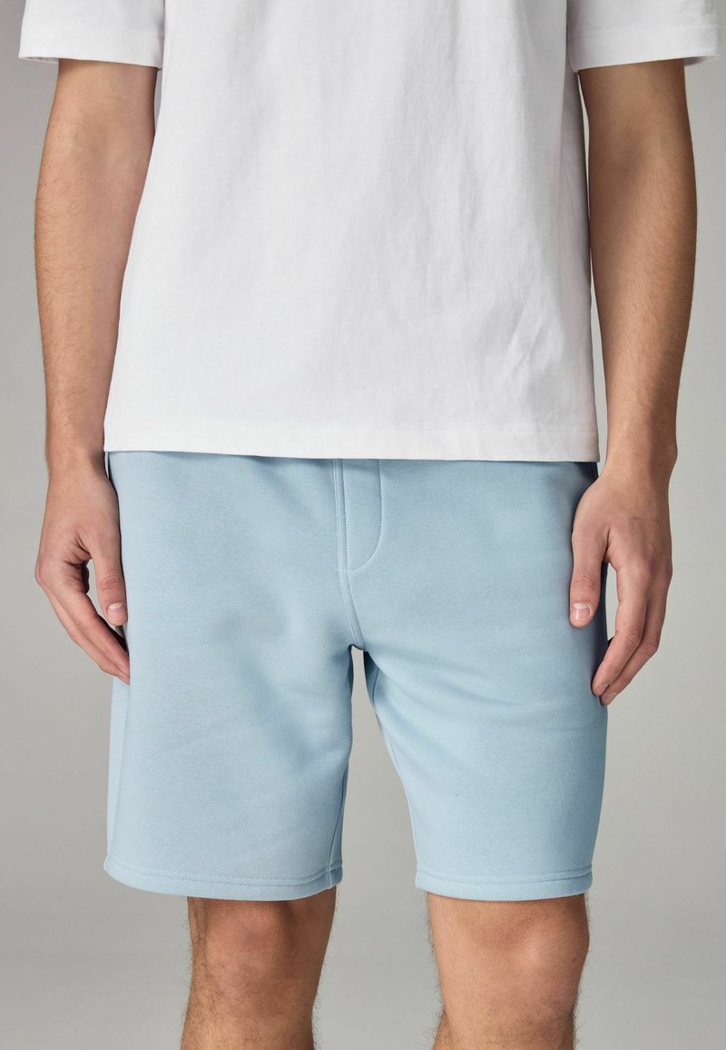 Pantaloni corti azzurri chiari realizzati in tessuto morbido, con una texture liscia, una forma aderente e una singola tasca frontale. Abbinati a una maglietta bianca.