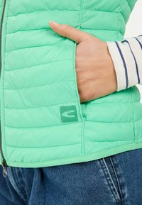 Hellgrüne gesteppte Weste mit Reißverschluss, Seitentasche und Logopatch. Kombiniert mit blauer Jeans und einem gestreiftenLangarmshirt.