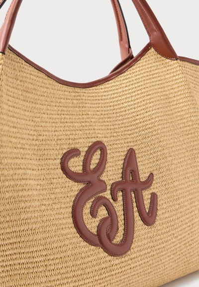 Πλεκτή τσάντα ώμου σε μπεζ χρώμα με καφέ δερμάτινες λαβές και μεγάλο διακοσμητικό διακοσμητικό λογότυπο από καφέ δέρμα στο μπροστινό μέρος.
