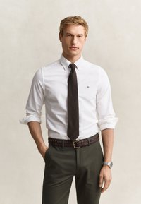 Homme avec manches blanches de chemise retroussées, cravate noire, ceinture tressée marron, pantalon foncé et montre argentée posant devant un fond clair.