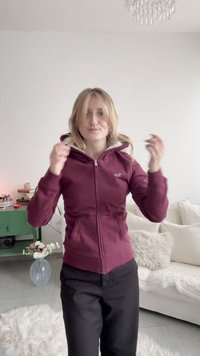 Kvinne i en burgunder hettejakke med glidelås som justerer håret i en lys stue med en hvit sofa og et grønt sidebord med blomster.