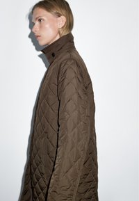 Bruine gewatteerde jas met een hoge kraag, voorzien van een ruitpatroon, knoopdetails en een gladde, glanzende textuur.